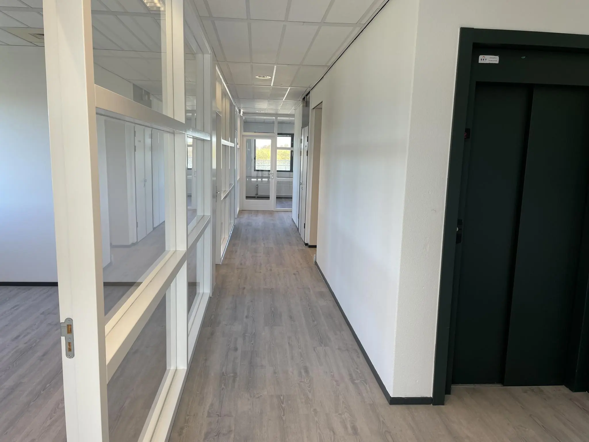 Moderne kantoorhal met houten vloer, witte wanden, glazen kantoorwanden en een donkere liftdeur aan de rechterzijde, gelegen aan de Laan van ’s-Gravenmade.